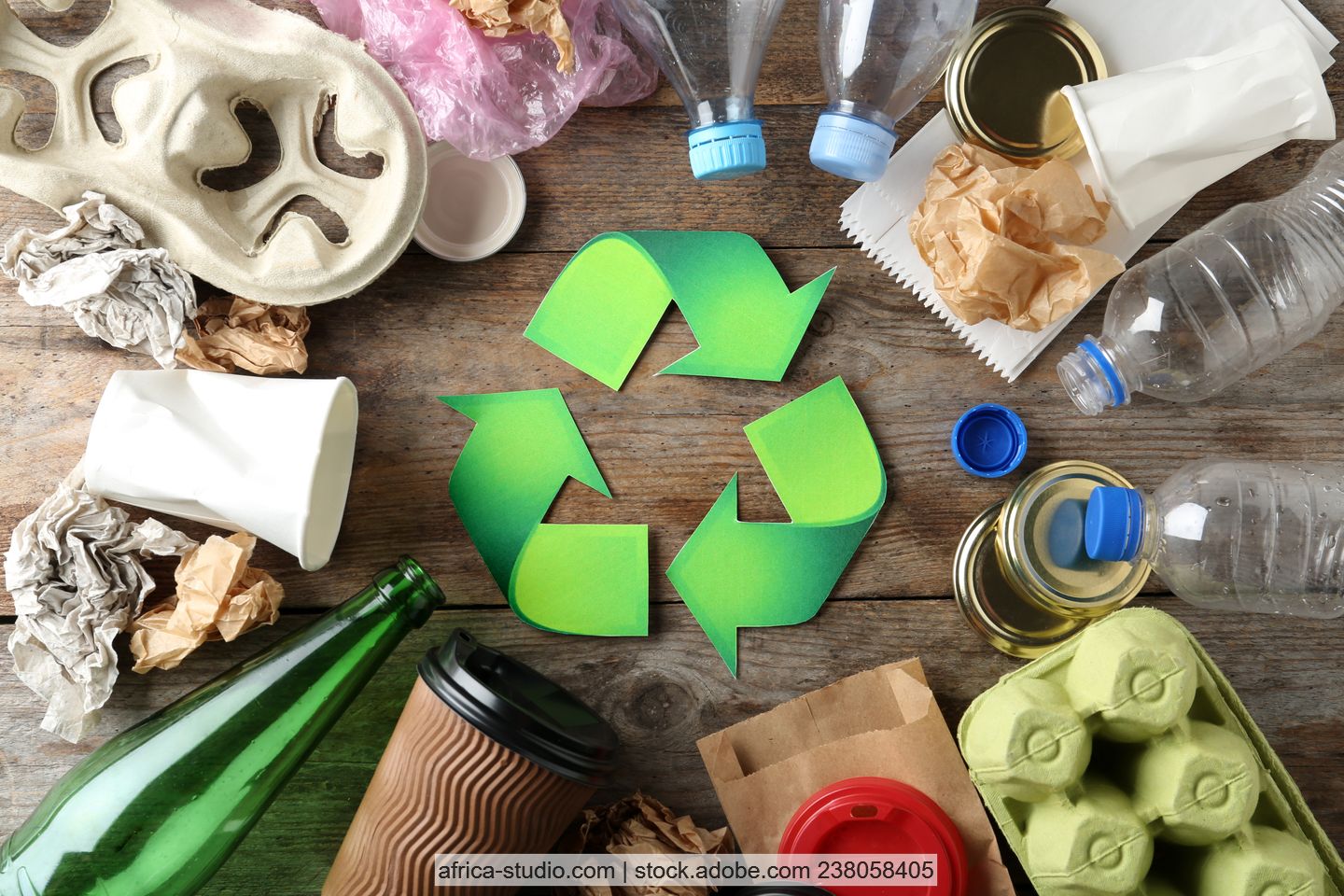 Verschiedene Verpackungen (Glas- und Kunststoffflaschen, Becher, Eierkarton, Metallverschlüsse von Gläsern) auf einer Holzfläche um ein grünes Recyclingsymbol herum gelegt.