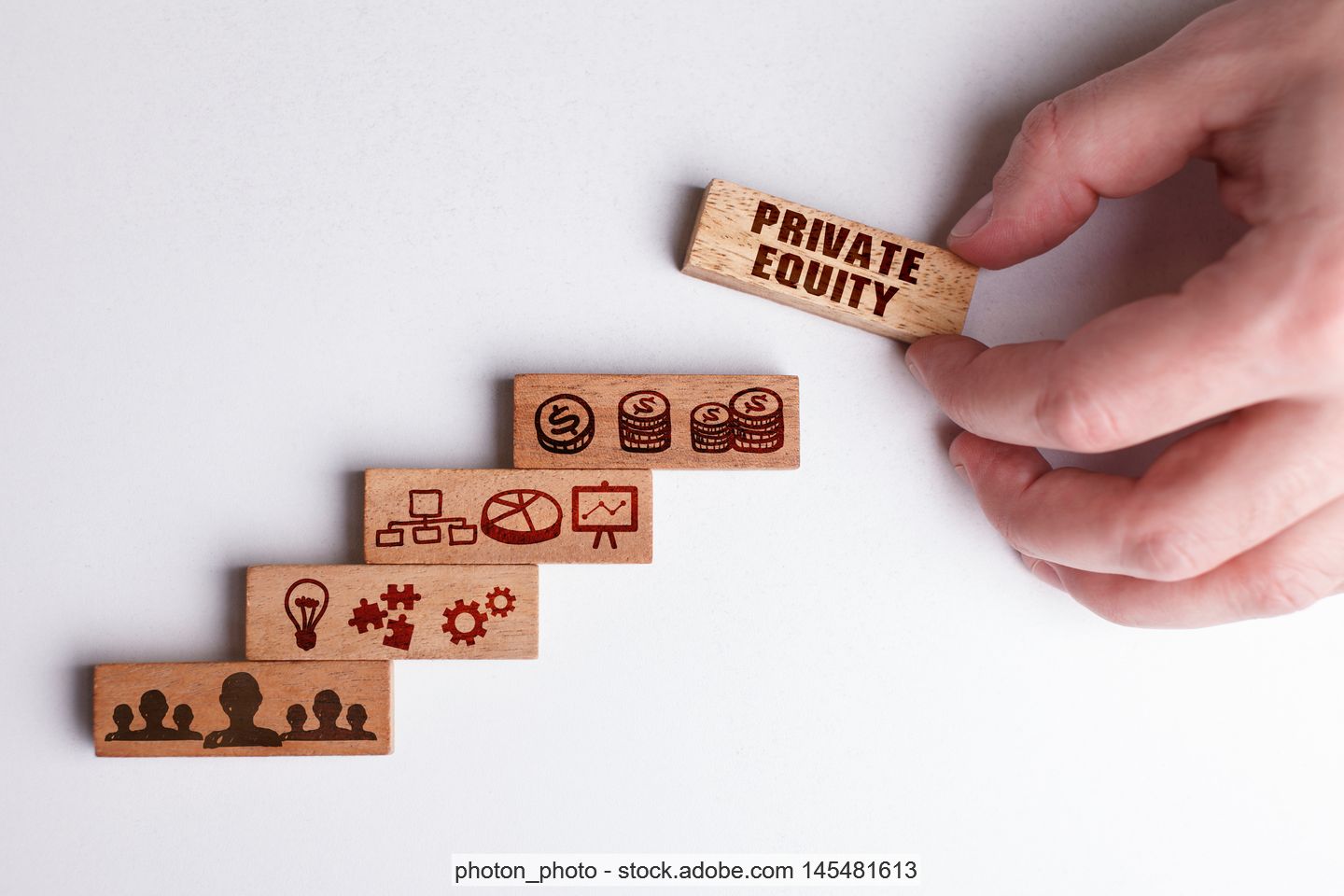 Hand auf Tisch und Holzstücke mit verschiedenen Symbolen von Köpfen, Münzen und Strukturen sowie der Aufschrift "Private Equity"